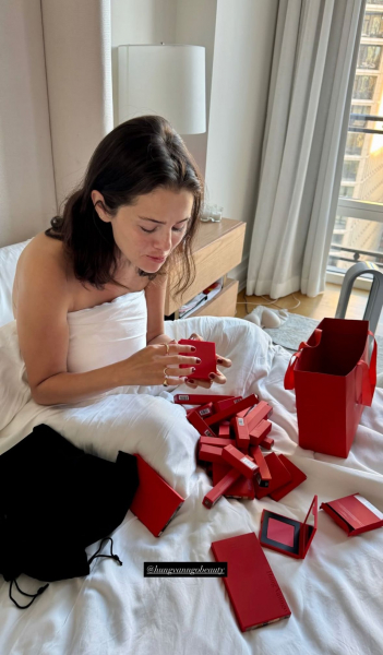 Selena Gomez wrapped in white bedding opening red makeup packaging